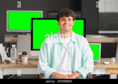 Smiling young man in a casual shirt sitting at a desk with Multiple green screen computer monitors in an office setting, Multiple computer monitors wi Stock Photo