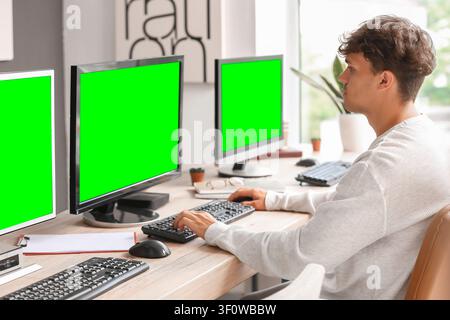 Young man working on multiple computers with green screen monitors at a modern home office desk, He is focused on his tasks, typing on a keyboard and Stock Photo