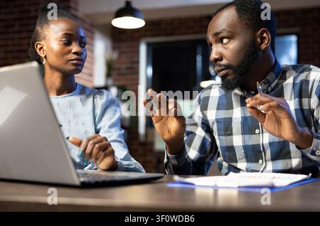 Colleagues analyzing forex market charts together Black male and female colleagues working late at night, analyzing forex market trends on a laptop and clipboard. Stock Photo