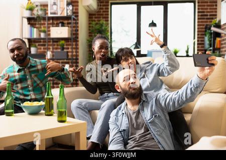 Happy diverse friends take selfies during relaxed weekend home gathering, joking around and capturing joyful memories. Multiethnic group enjoys drinks, laughter and friendly bonding. Stock Photo
