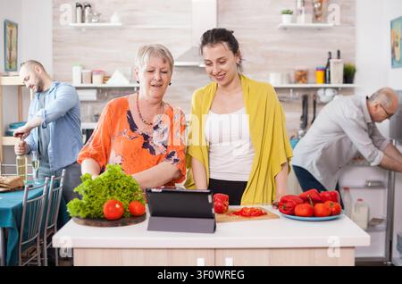 Mother and daughter following recipe on tablet, cheerful mother and daughter using tablet to cooking tasty salad following online recipe in kitchen Husband holding wine bottle Father looking inside. Stock Photo