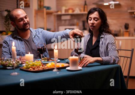 Young man pours red wine into his wife's glass while celebrating their relationship at a romantic dining table at home, adding romance, table setting and shared celebration detail. Stock Photo