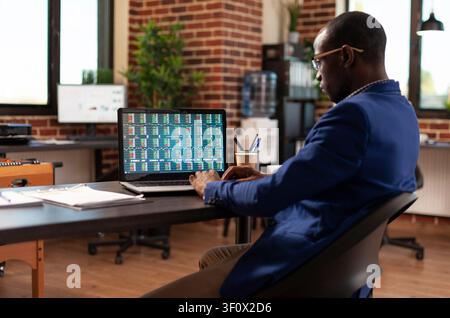 African American businessman studies market growth and investment charts on a laptop, analyzing trading data and global financial numbers in an office. Stock Photo