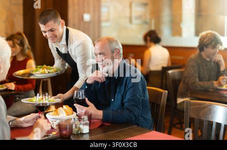 Young waiter bringing food to elderly couple in restaurant Stock Photo