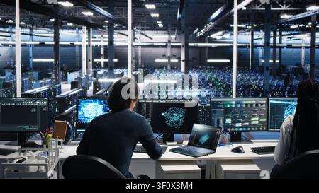 High tech server room IT technician arriving at the office, managing AI systems for uptime and efficiency, while IT professional arriving at work, sitting at desk, working with AI in computing hub. Stock Photo