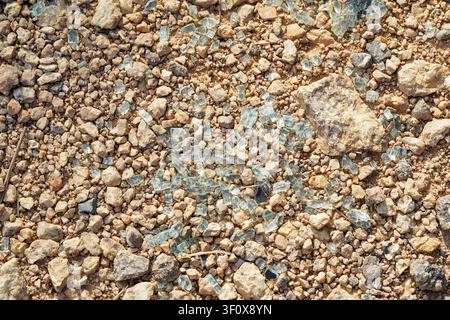 Close-up of numerous sharp, broken glass shards scattered among small beige and grey rocks and dirt, creating a hazardous, gritty texture. Full frame Stock Photo