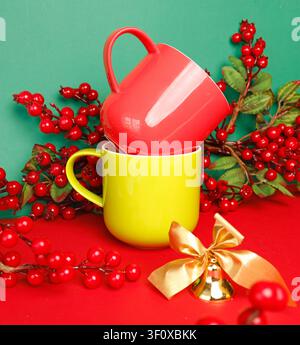 Christmas mockup red and yellow ceramic mugs stacked on red table against green background. New year bright cups template with winter holly berries Stock Photo