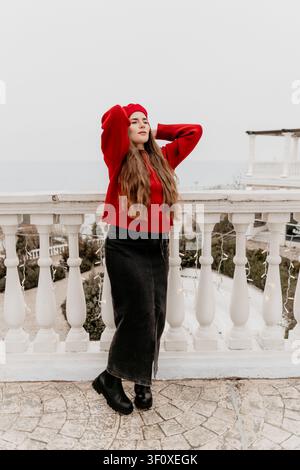 Woman balcony sea stylish young female in red sweater and beanie posing outdoors on white terrace with ocean view Stock Photo