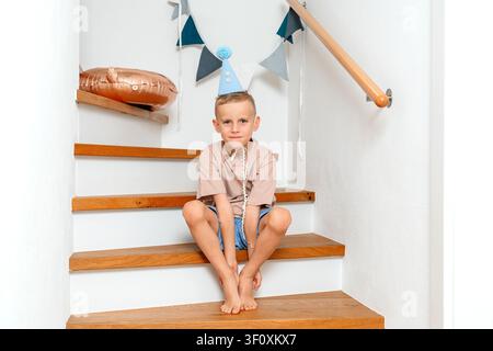 Boy sitting on stairs, in indoor staircase with white walls and wooden handrail, with staircase, wooden handrail, party hat, balloon, with wood Stock Photo