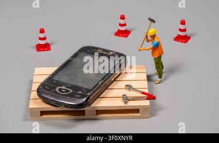 A small construction worker figure is using a hammer to fix a large smartphone placed on a wooden pallet. Traffic cones surround the area, suggesting Stock Photo
