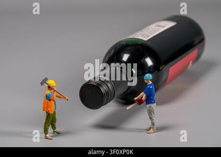 Two miniature construction workers stand near a giant wine bottle, engaged in conversation about their work. They hold tools, ready for their task. Stock Photo