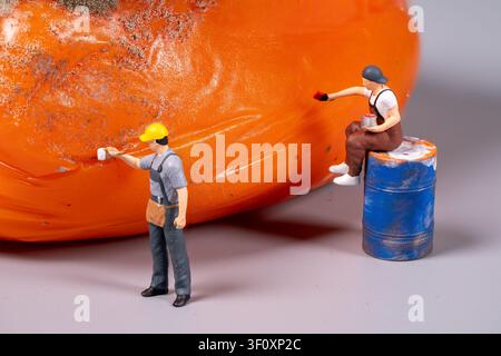 Two miniature workers are busy painting a large orange surface. One stands while applying paint, and the other sits on a barrel. Stock Photo