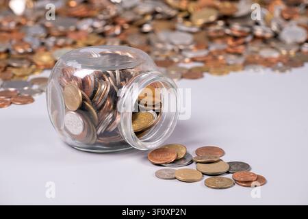 A glass jar filled with coins tips over, spilling various denominations onto a surface. Surrounding the jar are more scattered coins, showcasing diver Stock Photo