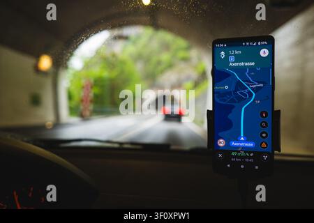 A smartphone mounted on a dashboard holder displays a navigation app route while driving out of an illuminated road tunnel with a car in front blurred Stock Photo