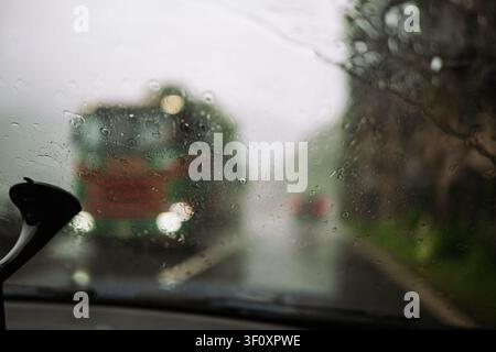 A point of view shot from inside a car looking through a wet windshield covered in raindrops during a storm, showing a blurry oncoming green truck wit Stock Photo