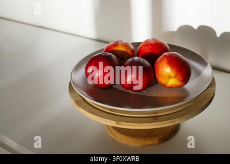 Ripe red nectarines arranged on a grey ceramic plate and a round bamboo serving stand. Clean minimalist composition with harsh shadows Stock Photo