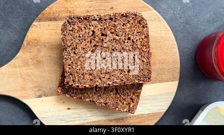Slices of dark seed bread placed on a wooden cutting board with a jar of jam and butter on a gray countertop  Stock Photo