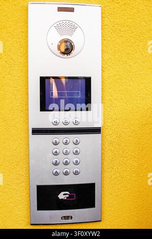 A contemporary silver outdoor intercom entry panel featuring a built-in camera, digital keypad, and RFID scanner on a textured yellow wall. Stock Photo