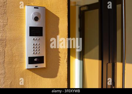 A contemporary silver outdoor intercom entry panel featuring a built-in camera, digital keypad, and RFID scanner on a textured yellow wall. Stock Photo