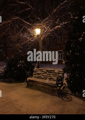 Snow-covered park bench under warm streetlight on winter night winter bench, snow covered bench, park bench, empty bench, winter Stock Photo