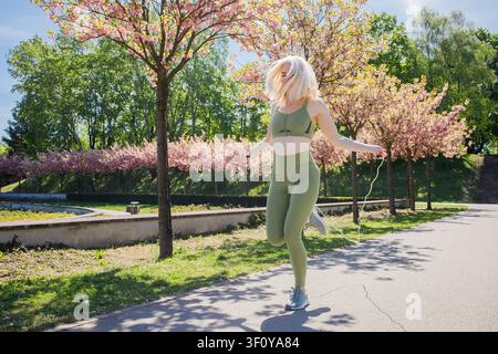 Blonde middleaged sporty woman jumping rope in spring sunny city park Stock Photo