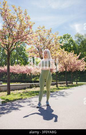 Blonde middleaged sporty woman jumping rope in spring sunny city park Stock Photo