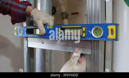 worker wearing gloves checks the accuracy of the installation of galvanized metal profiles using a blue spirit level. The frame of the future wall Stock Photo
