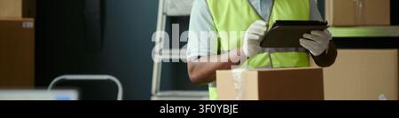 Header of worker wearing gloves checking inventory on tablet beside cardboard box, cropped view. Useful for warehouse logistics, stock control, fulfillment marketing Stock Photo