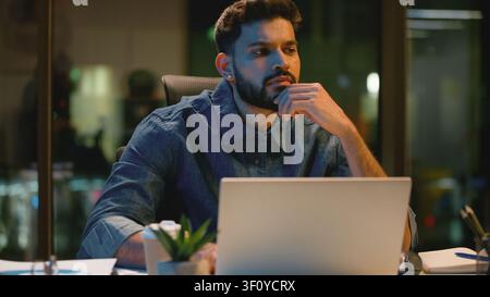 Pensive hesitate Indian business man with laptop thinking at dark office at night evening thoughtful serious Arabian male guy businessman Hindu worker Stock Photo