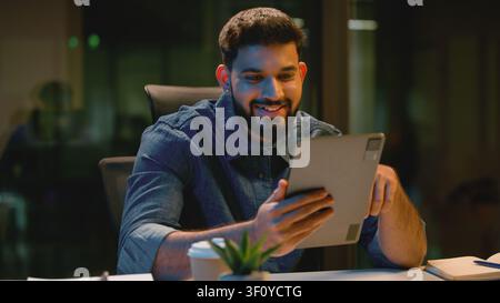 Smiling Indian business man Arabian businessman satisfied using digital tablet app at dark office at late time. Hindu male guy worker working using Stock Photo