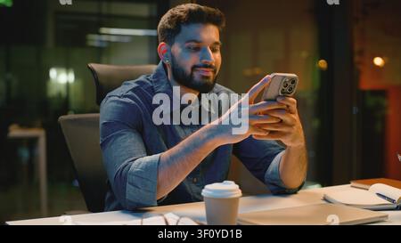 Happy Indian business man browsing mobile phone in evening office at table joyful Arabian businessman smiling scrolling web app Hindu male guy Stock Photo