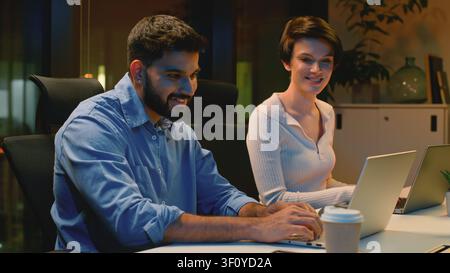 Two multiracial business colleagues working with laptops late at night evening dark office. Indian businessman with coffee cup work with Caucasian Stock Photo