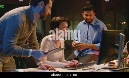 Diversity business team happy talking looking in computer screen at dark night office. Caucasian coworkers discuss online task Arabian and Caucasian Stock Photo