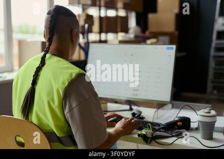 Rear view of young adult worker wearing safety vest, monitoring data on desktop computer in warehouse office. Suitable for logistics operations, inventory management marketing Stock Photo
