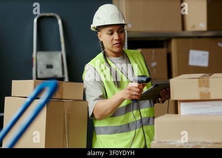 Medium shot of young adult biracial woman scanning packages with barcode reader and checking tablet in warehouse, managing inventory and fulfillment. Ideal for logistics, supply chain, ecommerce Stock Photo