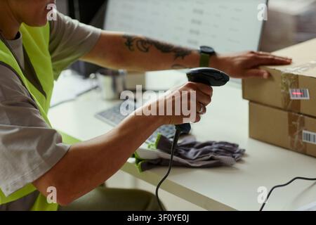 Close up of young adult warehouse worker scanning barcode on shipping boxes beside computer, processing orders for fulfillment and inventory use in e commerce logistics marketing Stock Photo
