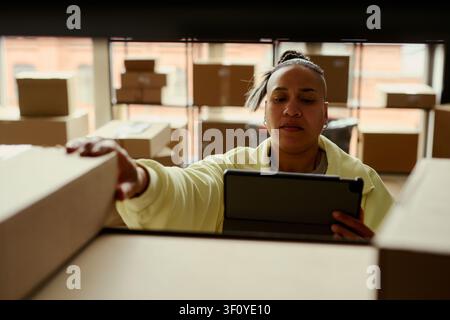Middle aged biracial woman checking inventory on tablet while reaching for shipping box in warehouse, supporting ecommerce fulfillment and small business logistics marketing Stock Photo