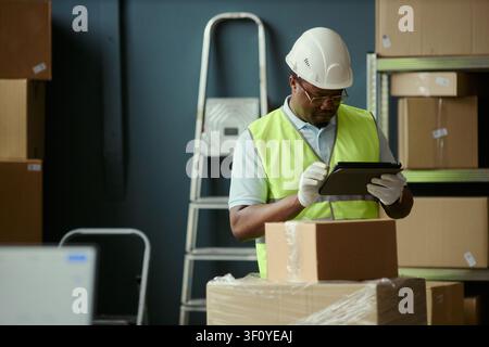 Medium shot of Black middle aged warehouse worker checking inventory on tablet beside stacked parcels, managing logistics workflow. Ideal for supply chain, fulfillment, shipping content Stock Photo