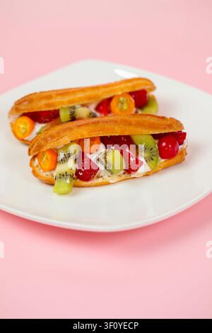 Two eclairs filled with fresh fruits sit on a white plate. The background is pink. The fruits include kiwi, strawberries, and cherries. Stock Photo