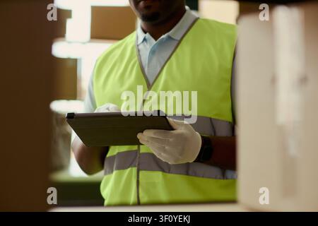 Medium shot of Black middle aged warehouse worker using tablet while checking inventory between shelves. Ideal for logistics, supply chain, fulfillment, stock control marketing Stock Photo