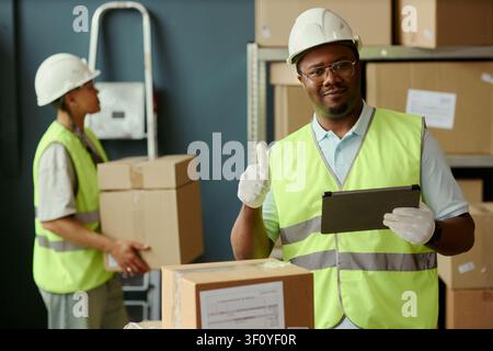 Portrait of Black young adult warehouse worker holding tablet and showing thumbs up while coworker carrying boxes in background, ideal for logistics staffing and delivery service marketing Stock Photo