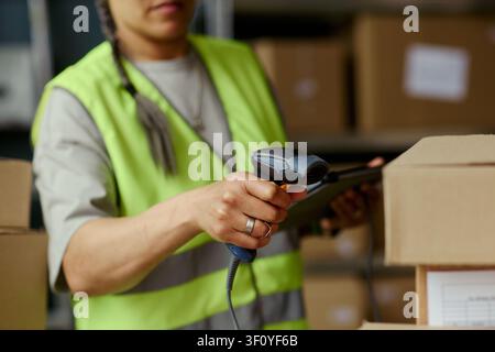 Young adult woman scanning barcode on shipping box while checking tablet in warehouse, managing inventory and order fulfillment. Useful for logistics, retail, supply chain marketing Stock Photo