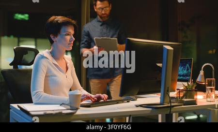 Caucasian leader business man with digital tablet intern woman using computer in dark night office boss company CEO near female worker browsing Stock Photo