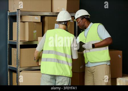 Medium shot of multiethnic warehouse workers checking inventory and handling shipping boxes beside storage racks. Suitable for logistics operations, supply chain, fulfillment marketing Stock Photo