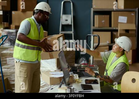African American man carrying parcel boxes while Asian woman scanning barcode in warehouse, managing inventory workflow. Useful for logistics, fulfillment, supply chain marketing Stock Photo