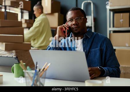 Medium shot of African American young adult man taking phone call while working on laptop in shipping office, managing orders and logistics. Ideal for ecommerce operations marketing Stock Photo