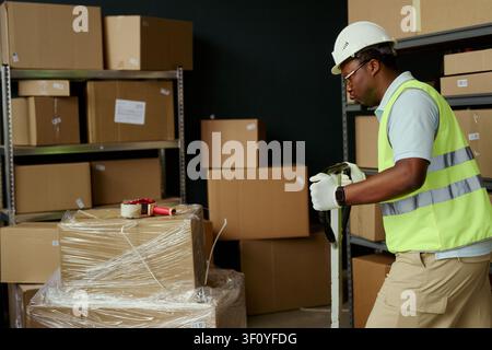Medium shot of African American young adult warehouse worker moving pallet jack beside wrapped boxes, handling logistics and inventory flow. Ideal for supply chain, shipping, fulfillment marketing Stock Photo