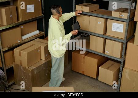 High angle view of young adult biracial woman checking inventory on digital tablet while reaching for parcel on warehouse shelf. Ideal for e commerce logistics, stock control, fulfillment marketing Stock Photo