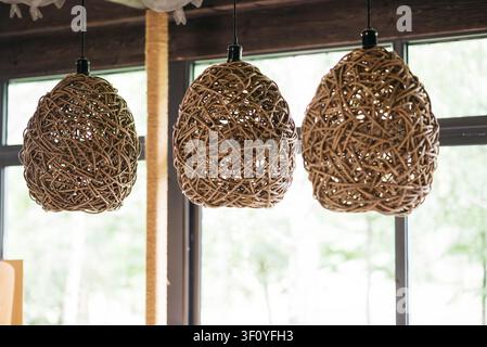 Three handmade wicker pendant lamps hanging in a cozy modern interior with large windows. Natural rattan lighting fixtures in Scandinavian, boho and eco style home decor. Soft daylight, sustainable design, rustic atmosphere. Stock Photo