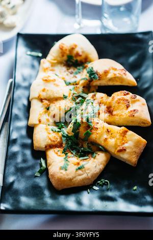 Freshly baked cheesy flatbread cut into slices and topped with chopped parsley, served on a black plate. Crispy golden crust, melted cheese create a delicious homemade appetizer perfect for restaurant menus, food blogs, and culinary themes Stock Photo